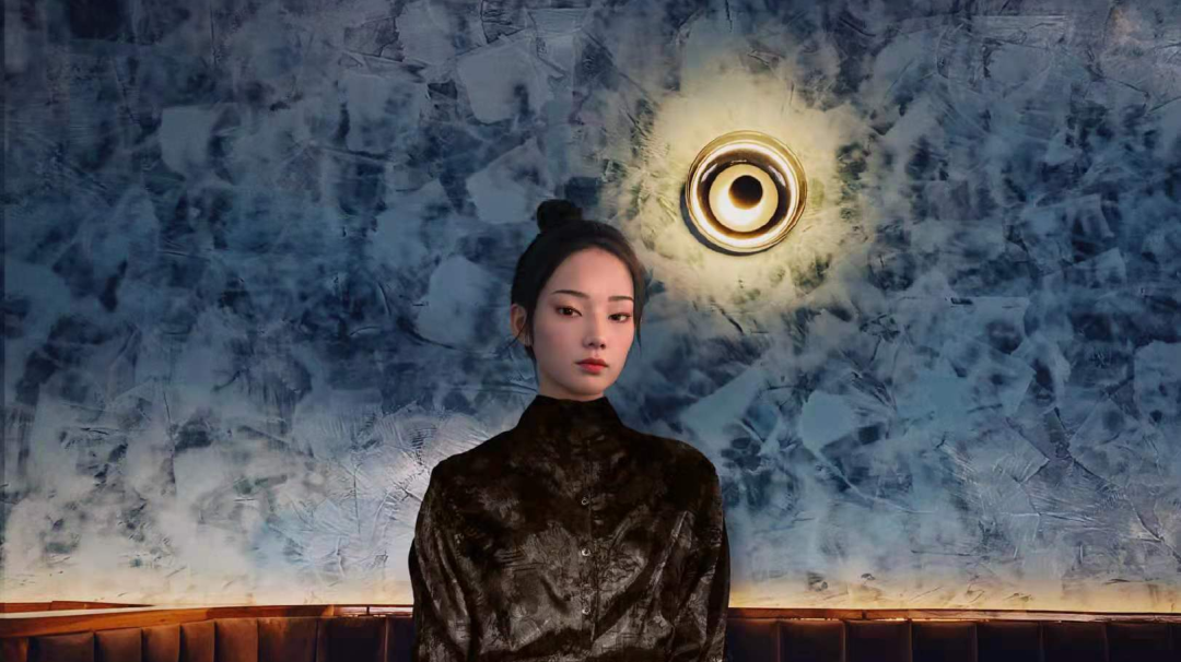 图片展示一位女性，穿着黑色上衣，背景是蓝色调的墙面和一个圆形的灯具。她的表情平静，发型是高束发髻。